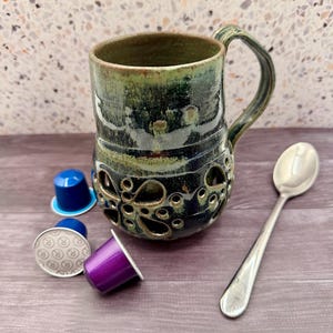 Puede incluir: Taza de cerámica hecha a mano con un diseño texturizado único en tonos verdes y azules. La taza presenta un asa decorativa y patrones recortados. Una cuchara plateada y varias cápsulas de café coloridas están junto a la taza.