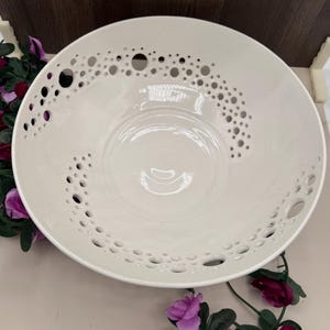 Puede incluir: Un cuenco de cerámica grande de color blanco roto con un interior liso y brillante, y un borde ancho decorado con una serie de recortes circulares de varios tamaños dispuestos en un patrón curvo a lo largo del borde superior.