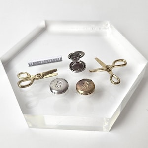 Puede incluir: Un juego de tijeras en miniatura de color dorado, una regla y dos pequeños polvos compactos con una letra 'B' en ellos. Los polvos compactos son uno plateado y uno dorado. Todos los artículos están en una bandeja hexagonal blanca.