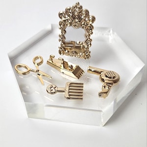 May include: Gold-toned hairdressing tools on a clear hexagonal display stand. The tools include a pair of scissors, a comb, a hair clipper, a hairdryer, and a decorative mirror.