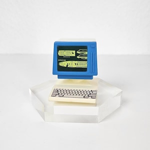 Könnte beinhalten: Ein Miniatur-Blau-Computer mit einem Bildschirm, der gelbe Grafiken anzeigt, und einer weißen Tastatur. Der Computer steht auf einer transparenten, geometrischen Basis und erzeugt eine Retro-Ästhetik.