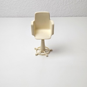 Könnte beinhalten: Ein weißer Drehstuhl aus Kunststoff mit einem runden Fuß und fünf Speichen. Der Stuhl ist für ein Puppenhaus oder eine Miniaturumgebung konzipiert.