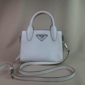 Może przedstawiać: Biała skórzana torebka z górnym uchwytem i odpinanym paskiem na ramię. Torba posiada srebrny metalowy zamek błyskawiczny i okucia. Logo Prada jest wyświetlane z przodu. Torba ma szerokość około 20 cm.