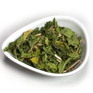 May include: A white, teardrop-shaped bowl filled with dried green mint leaves. The leaves vary in size and texture, with some appearing whole and others fragmented. The bowl sits on a white surface, creating a clean, simple presentation.