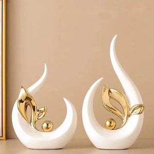 May include: Two white ceramic sculptures with gold leaf accents. Each sculpture features a stylised leaf design and a small gold sphere.