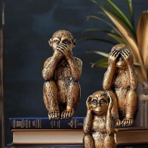 May include: Three gold monkey figurines, each with a different gesture: one covering its eyes, one covering its ears, and one covering its mouth. The monkeys are sitting on a stack of books.