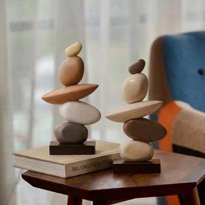 Pode incluir: Duas esculturas de pedra empilhadas, compostas por pedras ovais em tons de castanho e bege. As esculturas equilibram-se sobre bases castanhas escuras. O livro por baixo de uma escultura diz "BALANCING NATURE". Um elemento decorativo para a casa.