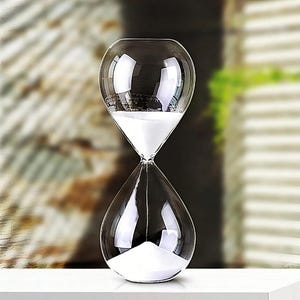 May include: A clear glass hourglass with white sand. The hourglass is on a white surface.
