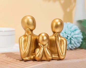 May include: Gold-toned abstract sculpture of a family, with two adults and a child, standing together. The sculpture is made of a smooth, shiny material and has a modern, minimalist design.