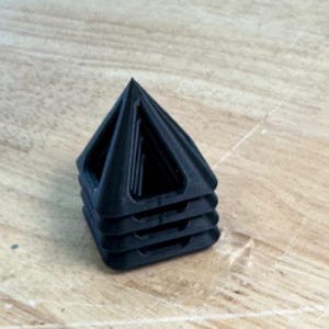 May include: A stack of four black, pyramid-shaped objects. Each pyramid has a hollow interior and a triangular base. The objects are made of a matte material and are stacked on top of each other. The background is a light brown wooden surface.