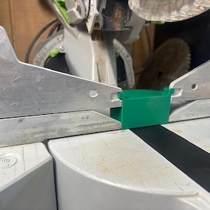 May include: Close-up of a miter saw with a green plastic component. The saw features silver metal blades and a white base. The image highlights the saw's design and construction, suitable for woodworking and carpentry tasks.