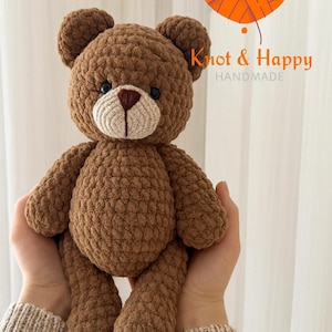 May include: A handmade brown crochet teddy bear with a beige muzzle and a heart-shaped nose. The bear has small black eyes and is held in someone's hands. The logo "Knot & Happy HANDMADE" is in the upper right corner.