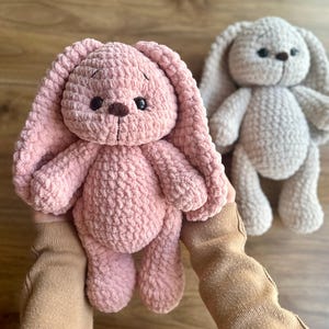 May include: Two handmade crochet bunny plushies. One is a soft pink, the other a light beige. Both have long floppy ears, small dark eyes, and a brown nose. The pink bunny is held, highlighting the plush texture. A charming, cuddly toy.