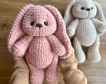 May include: Two handmade crochet bunny plushies. One is a soft pink color, and the other is a light beige. Both have long floppy ears, small dark eyes, and a brown nose. The pink bunny is held in a person's hands, showcasing the plush texture.
