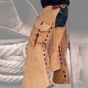 May include: Tan leather chaps with fringe and decorative stitching. The chaps have a pocket on the thigh and are secured with a belt. The fringe is a darker brown colour. The chaps are worn over blue jeans.