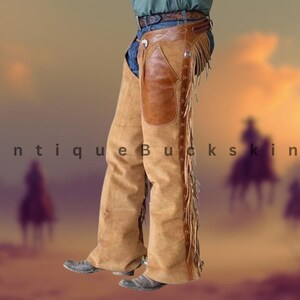 Op de afbeelding: Bruinleren chaps met franjes en een bruinleren patch op de dij. De chaps worden gedragen over een blauwe spijkerbroek en een bruine riem. De tekst "Antique Buckskin" is zichtbaar.