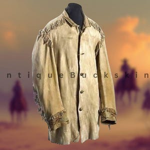 May include: A tan suede jacket with fringe detailing on the shoulders and sleeves. The jacket has a button-up front and a collar. The words "Antique Buckskin" are printed on the left side of the jacket. The background features a blurred image of riders on horseback.