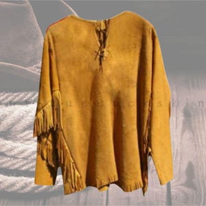 May include: Mustard-coloured suede shirt with long sleeves and fringe detailing on the sleeves. The shirt has a V-neck with a leather tie closure. The shirt is a rustic, western-style garment.