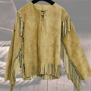 May include: A tan suede fringed shirt with long sleeves and a lace-up neckline. The shirt has fringe detailing on the sleeves and hem, giving it a Western or bohemian look. The garment is displayed on a black hanger.