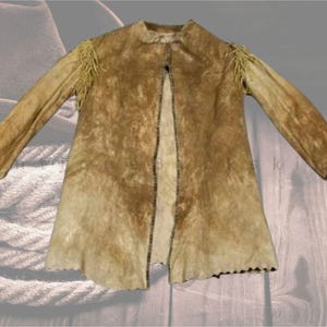 May include: A tan suede jacket with long sleeves and fringed shoulder detail. The jacket has a button closure and a scalloped hemline. The garment appears vintage or handmade, with a rustic aesthetic. The jacket is laid flat, showcasing its design.