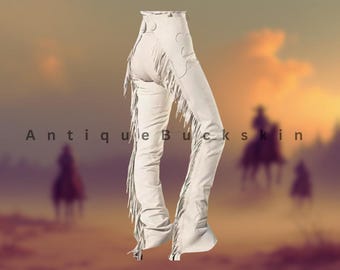 May include: Cream-colored leather chaps with decorative cutouts and long fringe detailing. The chaps are high-waisted and feature a classic Western design. The text "Antique Buckskin" is visible in the image.