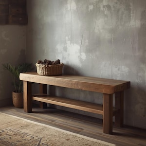 Op de afbeelding: Een massieve houten bank in warmbruine kleur met een onderste plank. Op de bank staat een gevlochten mand gevuld met dennenappels. Het hout heeft een natuurlijke nerf. Links staat een plant in een pot.