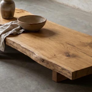 May include: A low-profile rectangular coffee table crafted from a single thick slab of light brown natural wood with a live edge. The table features a visible wood grain, knots, and two sturdy wooden block supports. A ceramic bowl and vase sit on top.