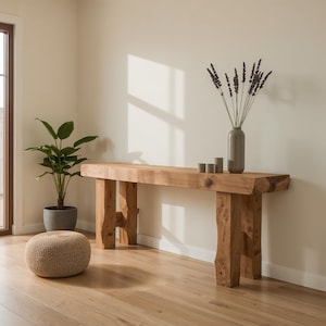 Può includere: Una consolle in legno rustico con finitura naturale. Il tavolo è sostenuto da gambe spesse e robuste. Un vaso grigio con fiori secchi e tre piccole candele sono sul tavolo. Una pianta in vaso e un pouf intrecciato sono in primo piano.