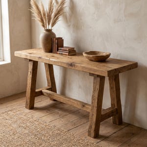 Puede incluir: Una mesa consola rectangular de madera con acabado natural y veta visible, con cuatro patas robustas conectadas por un travesaño horizontal. Encima hay un jarrón de cerámica marrón con hierba de la pampa seca, tres libros apilados y un cuenco de madera.