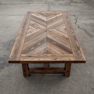 Może przedstawiać: Prostokątny drewniany stolik kawowy z wzorem w jodełkę na blacie. Stół wykonany jest z jasnego i ciemnobrązowego drewna i ma solidny, rustykalny design. Stół ma około 152 cm długości i 76 cm szerokości.