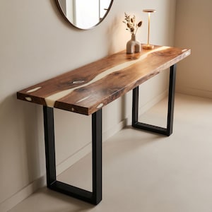 Peut inclure: Une console rectangulaire dotée d'un plateau en bois naturel avec une incrustation centrale en résine blanche suivant le grain. La table est soutenue par deux pieds en métal noir en forme de U. La surface en bois présente des nœuds visibles.