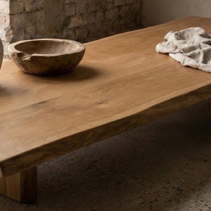 May include: A low-profile rectangular wooden coffee table with a natural live edge, featuring a smooth light brown grain. On the surface sits a beige ceramic vase with thin branches, a round wooden bowl, and a draped piece of beige linen fabric.