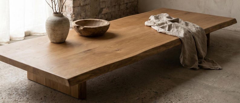 May include: A low-profile rectangular wooden coffee table with a natural live edge, featuring a smooth light brown grain. On the surface sits a beige ceramic vase with thin branches, a round wooden bowl, and a draped piece of beige linen fabric.