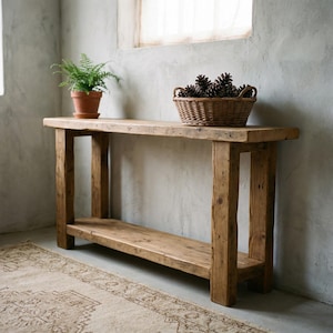 May include: A rustic wooden console table with a lower shelf and four sturdy legs. A small potted fern sits on the left, and a woven basket filled with pine cones rests on the right. The table is made of light brown wood.