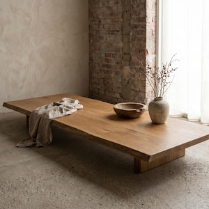 May include: A low-profile rectangular wooden coffee table with a natural live edge, featuring a light brown wood grain finish. A beige linen cloth is draped on the left side, while a wooden bowl and a ceramic vase with branches sit on the right.