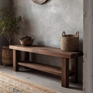 Op de afbeelding: Een donkerbruine houten bank met een onderste plank. Een gevlochten mand met dennenappels staat op de bank, samen met een gevlochten mand met handvatten. Een potplant en een decoratief tapijt zijn ook zichtbaar.