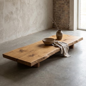 May include: A low-profile rectangular coffee table crafted from a single thick slab of natural light brown wood with visible grain and knots. The table features a live edge, a ceramic bowl, a small vase with dried branches, and a beige linen cloth.