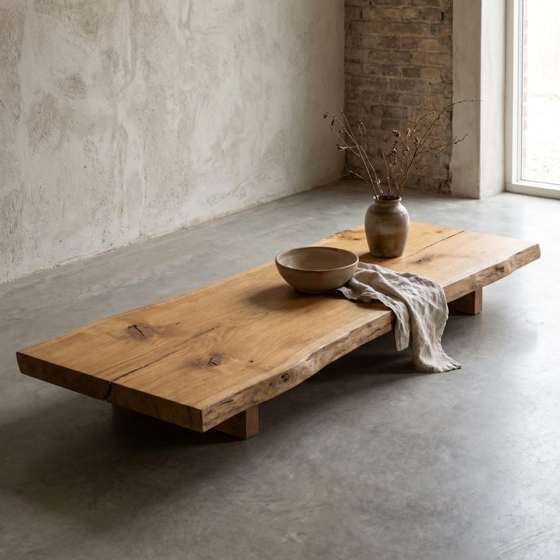 May include: A low-profile rectangular coffee table crafted from a single thick slab of natural light brown wood with visible grain and knots. The table features a live edge, a ceramic bowl, a small vase with dried branches, and a beige linen cloth.
