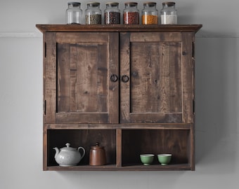 May include: A dark brown wooden wall cabinet with two doors featuring circular metal pulls. The top surface holds six clear glass jars filled with various grains and legumes. The open lower shelf contains a light gray teapot, a brown jar, and two small green bowls.
