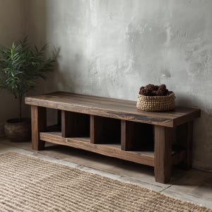 Peut inclure: Banc en bois rustique de couleur marron foncé. Il possède une étagère inférieure avec trois compartiments ouverts. Un panier en osier rempli de pommes de pin est posé sur le banc. Une plante en pot est visible à gauche.