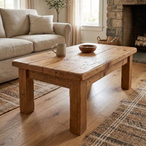 May include: A rectangular coffee table constructed from light brown wood with a visible grain and plank-style surface. The table features four sturdy, thick legs and a smooth finish. A single ceramic mug and a small wooden bowl sit on the top.