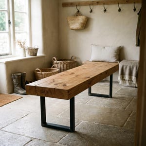 May include: A rectangular wooden bench featuring a thick, natural-toned timber seat with visible grain and knots, supported by two black metal U-shaped legs. The bench is positioned horizontally in a room with stone flooring and wicker baskets.