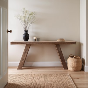 May include: A rustic wooden console table with angled legs, holding a black vase with white flowers, two candles, and a woven basket. A woven rug sits on the floor. The table is approximately 72 inches long.