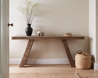 Pode incluir: Uma mesa consola retangular de madeira com pernas angulares, apresentando um vaso preto com ramos finos, duas pequenas velas cilíndricas e um cesto de vime colocado no chão. A mesa tem um acabamento de grão de madeira natural e construção sólida.