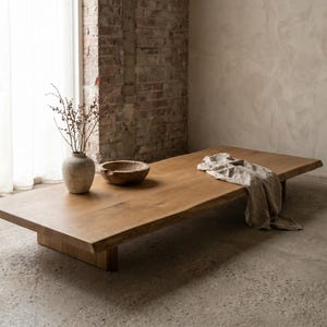 Peut inclure: Une table basse rectangulaire en bois à profil bas avec un bord naturel, présentant une finition en grain de bois brun clair. Sur la surface se trouvent un vase en céramique beige avec des branches fines, un petit bol en bois et un tissu en lin beige drapé.