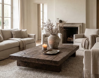 Puede incluir: Una mesa de centro baja y rectangular construida con gruesos tablones de madera oscura recuperada. La mesa presenta una base robusta en forma de bloque y una superficie desgastada y texturizada. Un jarrón de cerámica claro y redondo descansa sobre ella.