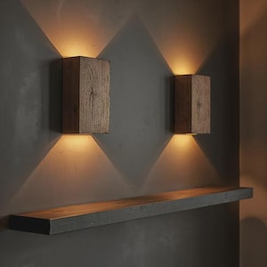 Può includere: Due applique da parete in legno con illuminazione calda, montate su un muro grigio. Le applique hanno una forma rettangolare e una finitura a venatura naturale. Una mensola in legno abbinata è montata sotto.