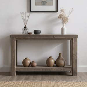 Könnte beinhalten: Ein rustikaler Holztisch mit unterem Regal, der verschiedene dekorative Gegenstände zeigt. Auf der Tischplatte befinden sich ein Glasdiffusor, eine kleine Schale und eine schwarze Keramikschale. Eine weiße Vase mit Trockenblumen und mehrere Holzvasen befinden sich auf dem unteren Regal.