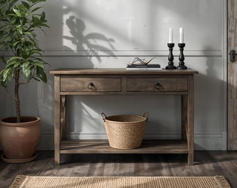 May include: A rectangular wooden console table with two drawers featuring dark circular pulls. The table has a lower shelf holding a woven seagrass basket. On the tabletop sit two black candle holders with white candles and a stack of books.