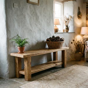 Könnte beinhalten: Rustikale Holzbank mit unterem Regal, vor einer strukturierten grauen Wand. Ein Weidenkorb mit Tannenzapfen steht auf der Bank. Eine Topffarn und ein gemusterter Teppich ergänzen das gemütliche Interieur.